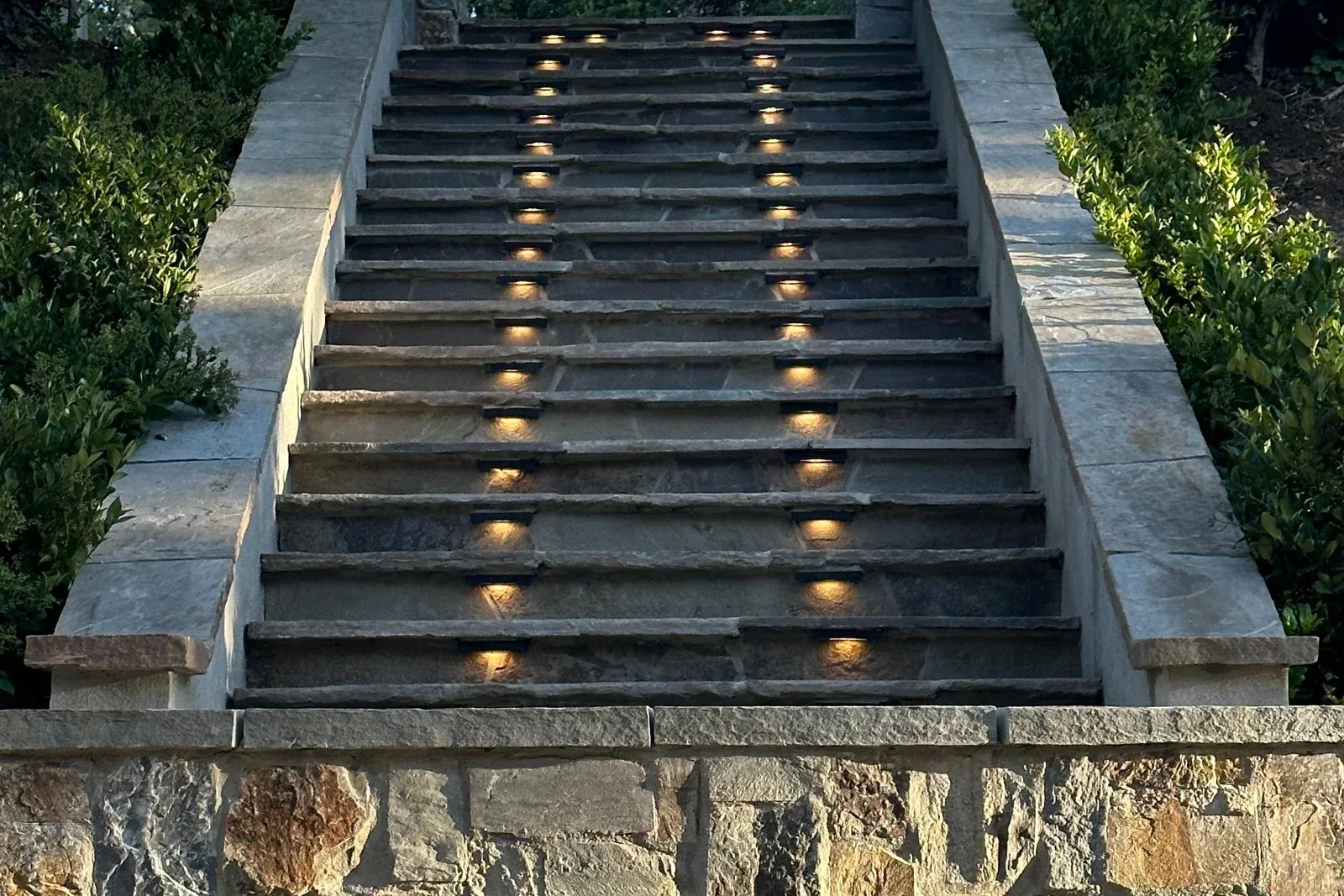 Illuminated stone stairs between capped walls and clipped hedges at dusk.